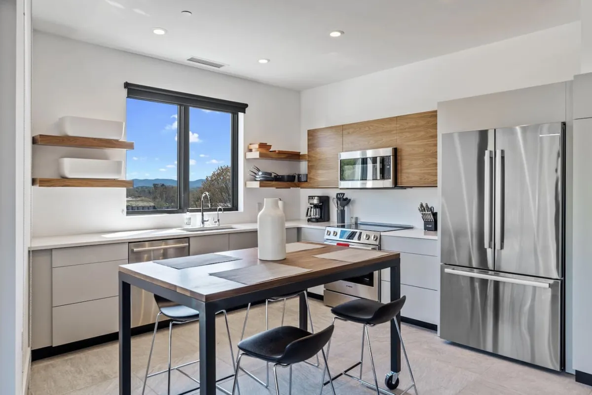 An open dining area with a central table and large window framing city views. Wooden shelves, stainless steel appliances, and contemporary decor create a warm, inviting space ideal for sharing meals and entertaining guests.