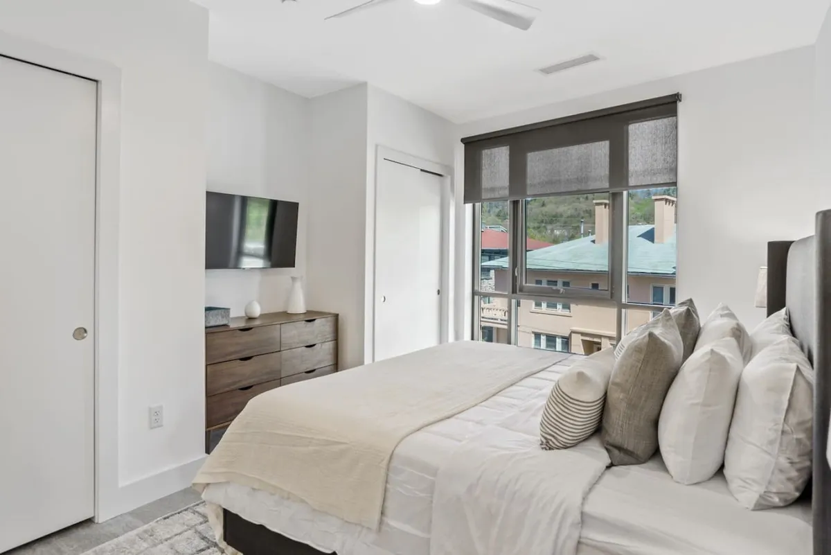 A serene bedroom featuring a large bed with decorative pillows, a wall-mounted TV, and a spacious window that fills the room with natural light. The overall atmosphere is tranquil and inviting, ideal for restful nights or cozy mornings.