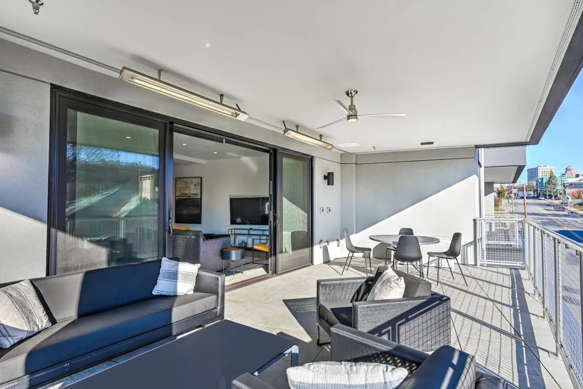 This image showcases an inviting outdoor seating area connected to the condo by sliding glass doors, with a view of the surrounding area. The setup includes comfortable seating and hints of social activity, blending indoor comfort with outdoor adventure. The surrounding cityscape visible beyond the balcony speaks to the property's proximity to Asheville's vibrant downtown.