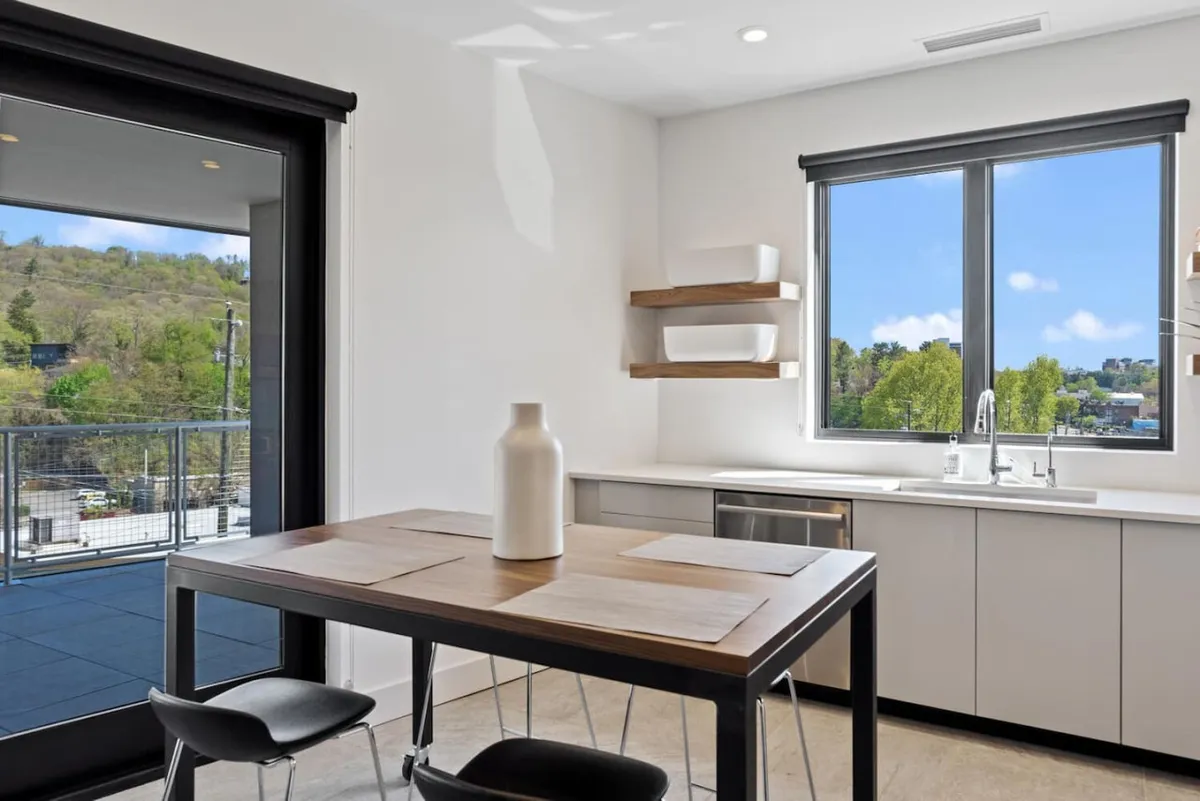 A bright and inviting dining area with a central dining table set near a window offering city views. Shelves lined with dishes add a homey feel, and the natural light streaming in enhances the mood for meals or group activities.