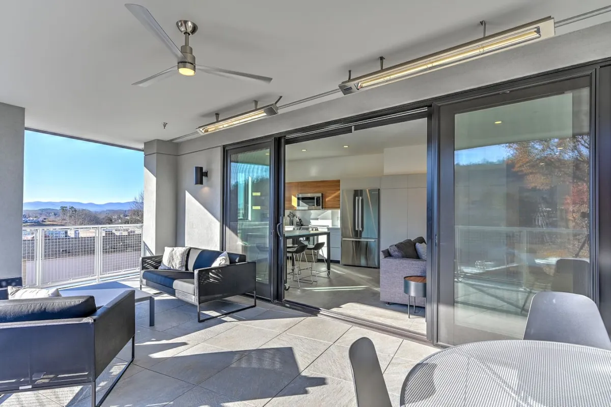 The image showcases the condo’s stylish outdoor seating area on a heated balcony, overlooking Asheville’s vibrant downtown and rolling landscape. The space is bathed in natural light, framed by contemporary furnishings, and seamlessly connects to the inviting interior living area, suggesting a perfect spot for sunrise breakfasts or evening gatherings.