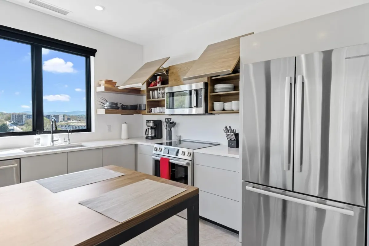 This image features a contemporary kitchen with a large window that lets in natural light, a spacious kitchen island, sleek stainless steel appliances, and open shelving. The layout communicates a space designed for both cooking and socializing, with ample room for preparing meals or gathering around the island.