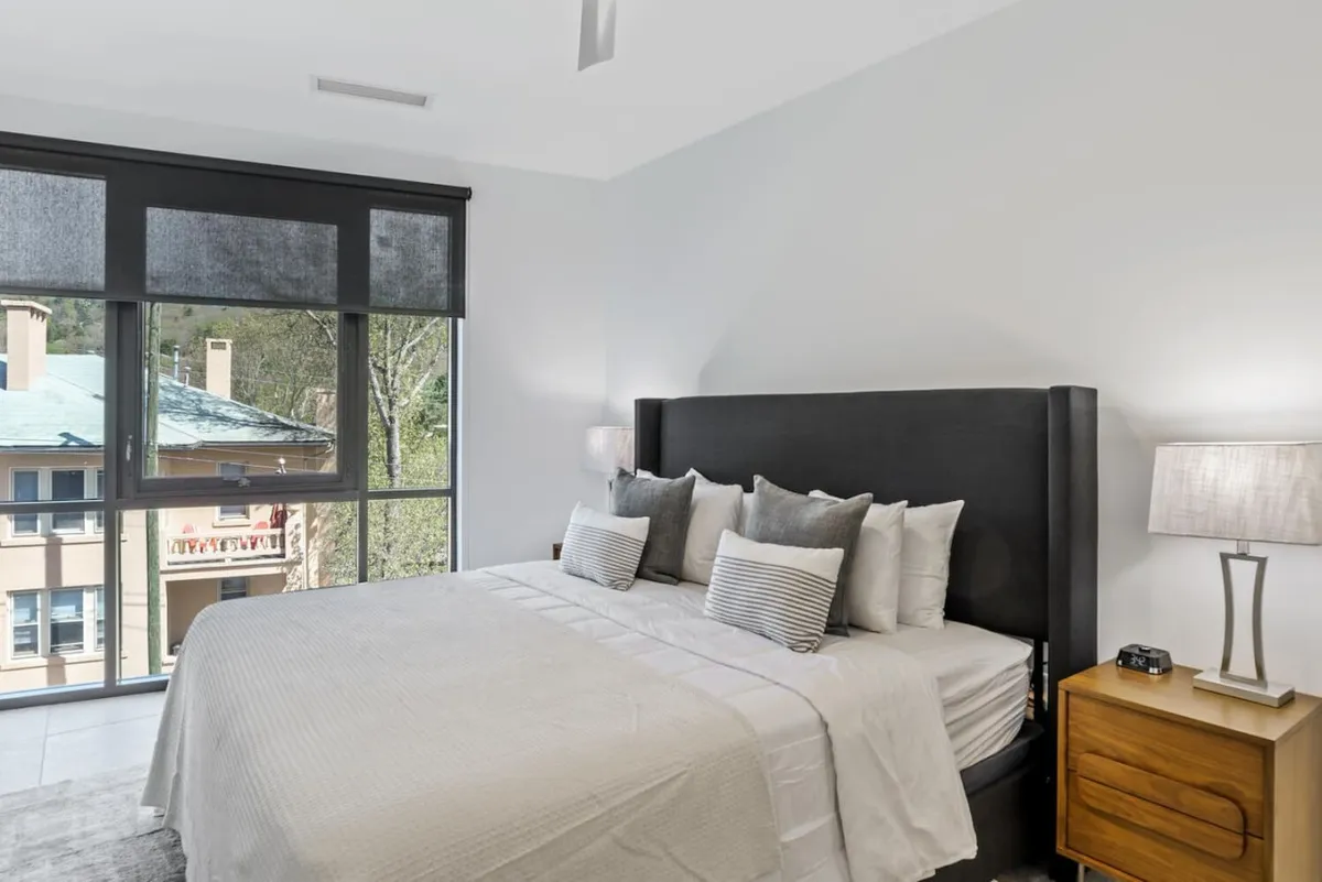A tranquil bedroom scene with a cozy bed adorned with decorative pillows, a nightstand with a lamp, and a window offering light and a welcoming view. The space is ideal for relaxation or quiet moments.