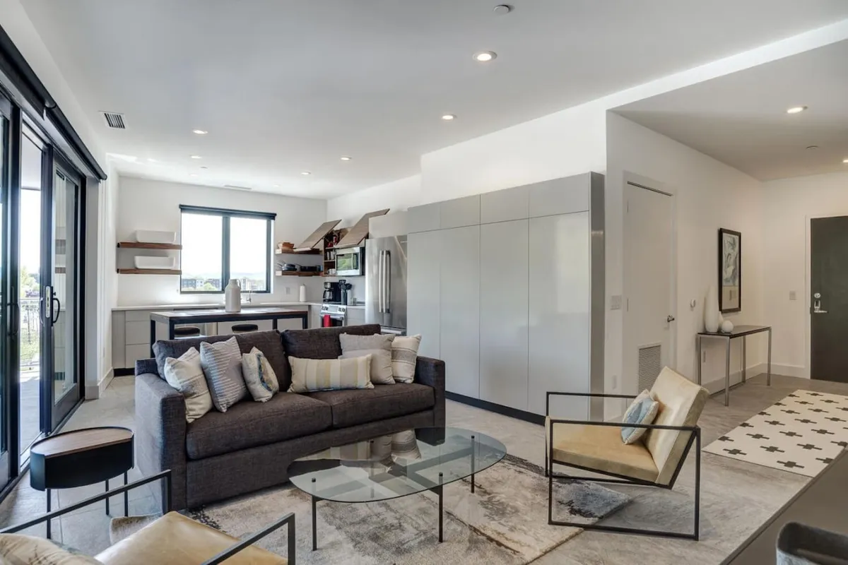 A contemporary living room featuring a cozy sofa and coffee table, bathed in natural light from large windows that offer a view of the city. The space is designed for socializing and relaxation, with decor that feels both stylish and inviting.